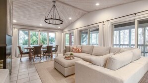 A spacious living room with a large beige sectional sofa, a dining area with a wooden table and chairs, and large windows offering a view of the outdoors. - Glennhouse Getaway (South Haven)