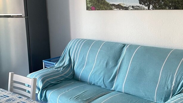 A room with a sofa, a table with chairs, a refrigerator, and a painting of a coastal landscape. - 2 Bedroom Apartment in Villasimius (Villasimius)