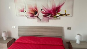 A bedroom with a wooden headboard, a red bedspread, and a wall-mounted floral artwork. - 2 Bedroom Apartment in Villasimius (Villasimius)
