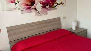 A bedroom with a wooden headboard, a red bedspread, a nightstand with a lamp, and a floral painting on the wall. - 2 Bedroom Apartment in Villasimius (Villasimius)