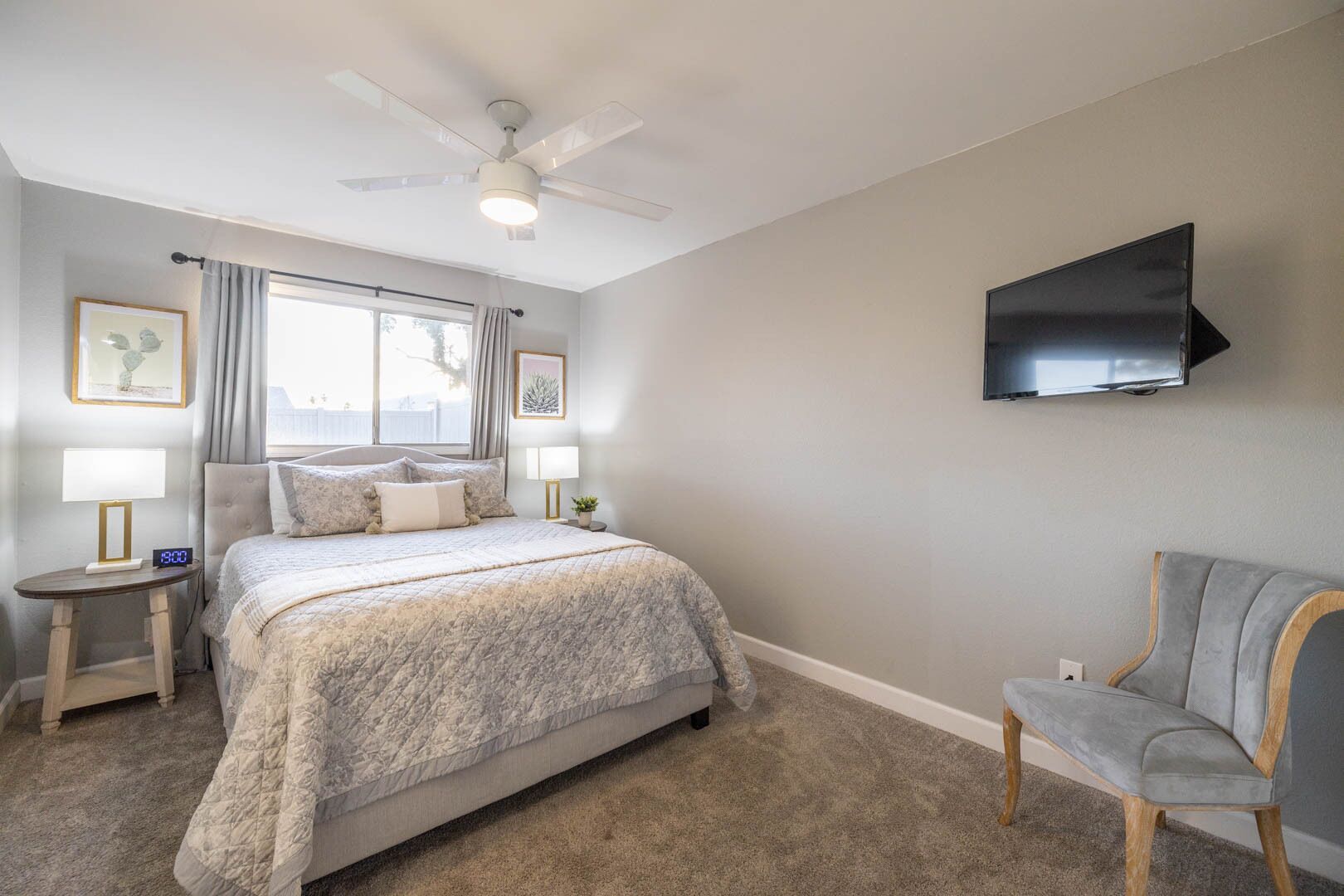Una camera da letto con un letto, comodini, una sedia, una ventola a soffitto, una televisione e una finestra con tende.