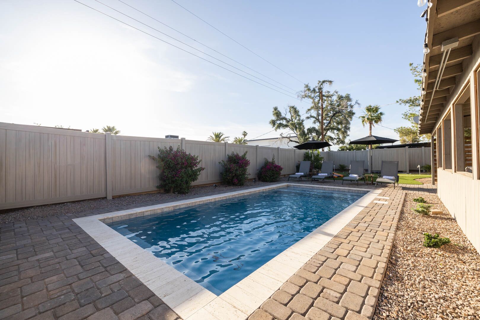 A rectangular swimming pool with a tiled edge, surrounded by a brick patio. There are lounge chairs and a covered area with umbrellas.