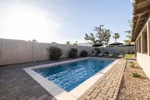 Une piscine rectangulaire dont le bord est recouvert de carreaux, entourée d’une terrasse en briques. On y trouve des chaises longues et un espace abrité avec des parasols.
