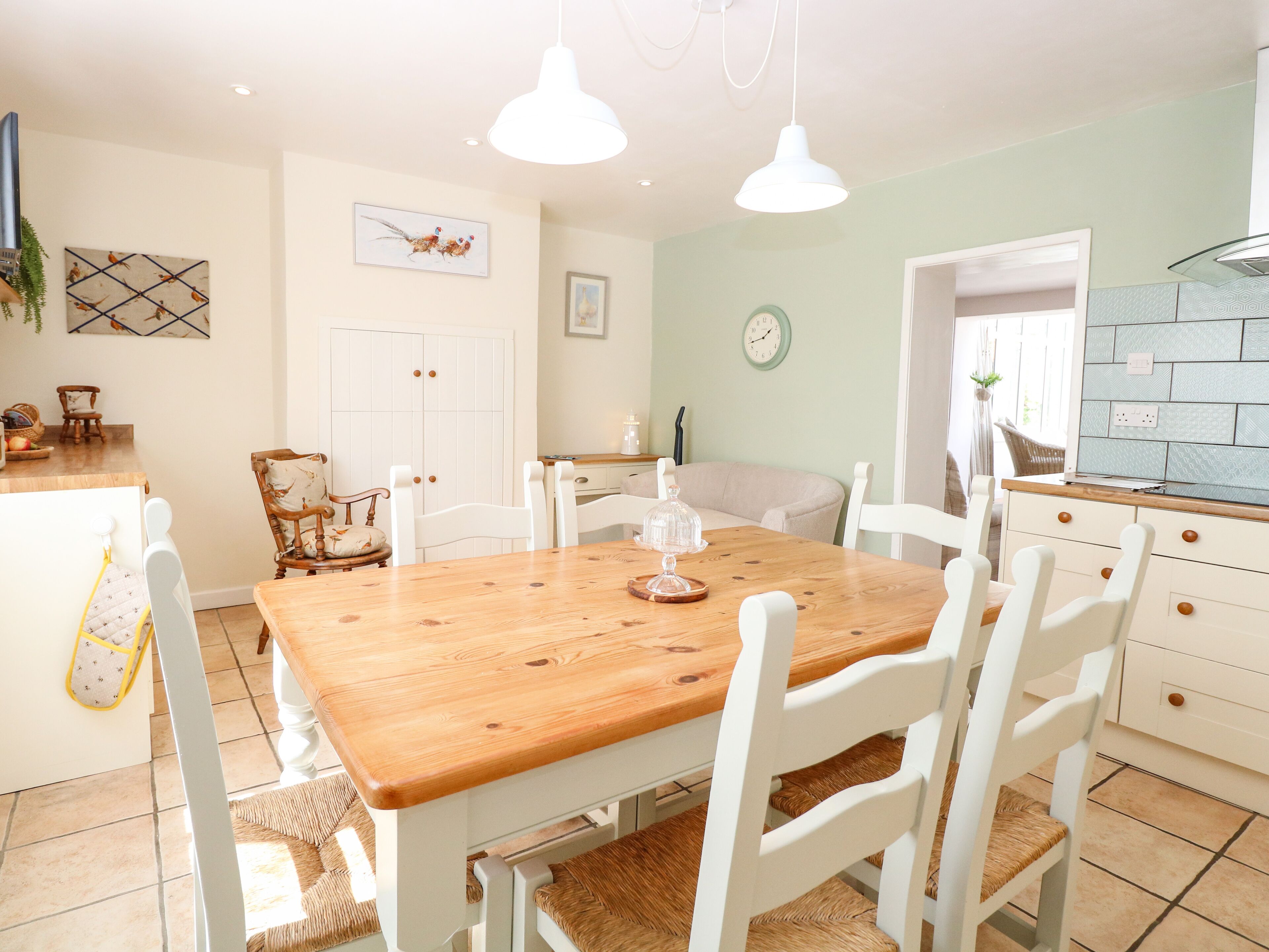 A dining area with a wooden table and white chairs, a kitchen with a blue backsplash, and a clock on the wall.