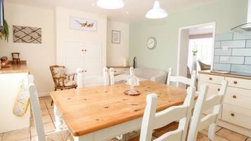 A dining area with a wooden table and white chairs, a kitchen with a blue backsplash, and a clock on the wall.