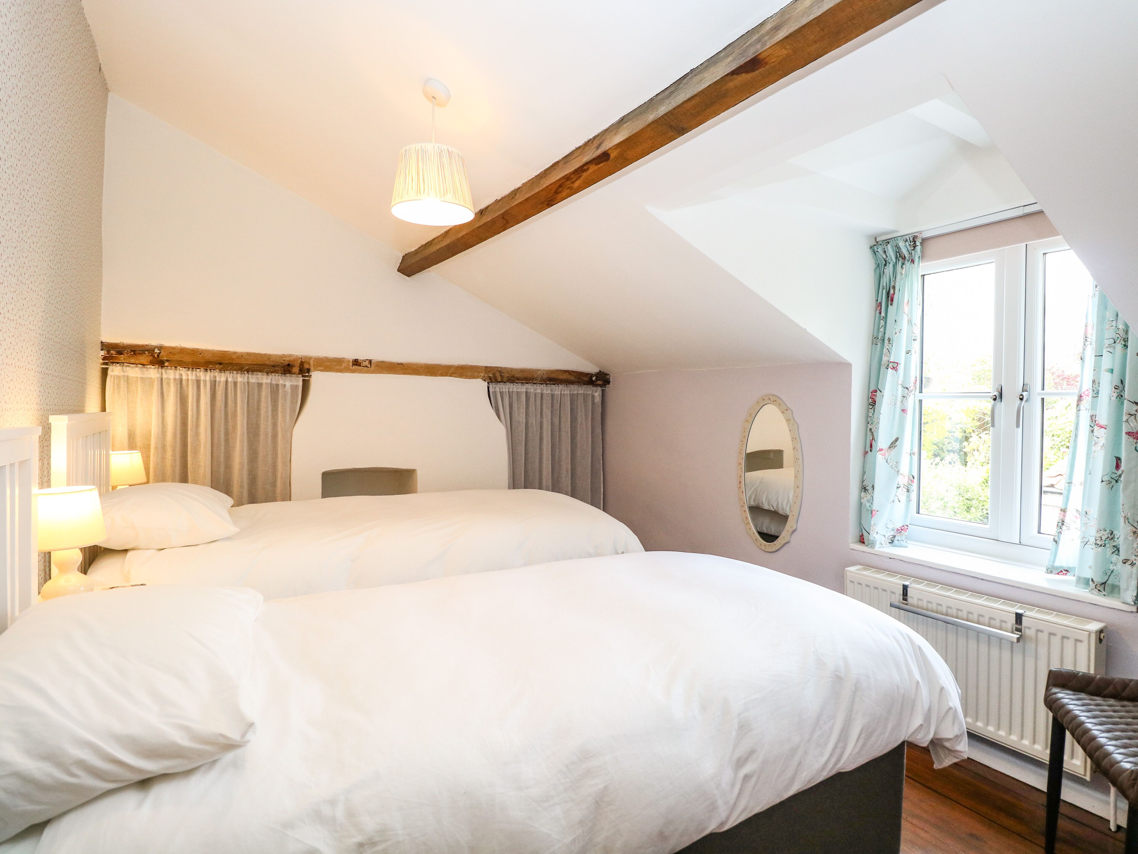 A bedroom with two beds, a window with curtains, a radiator, and a wooden ceiling beam.