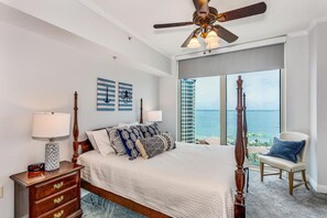 Quarto com cama grande, ventoinha no teto, vista para o oceano e cadeira perto da janela.