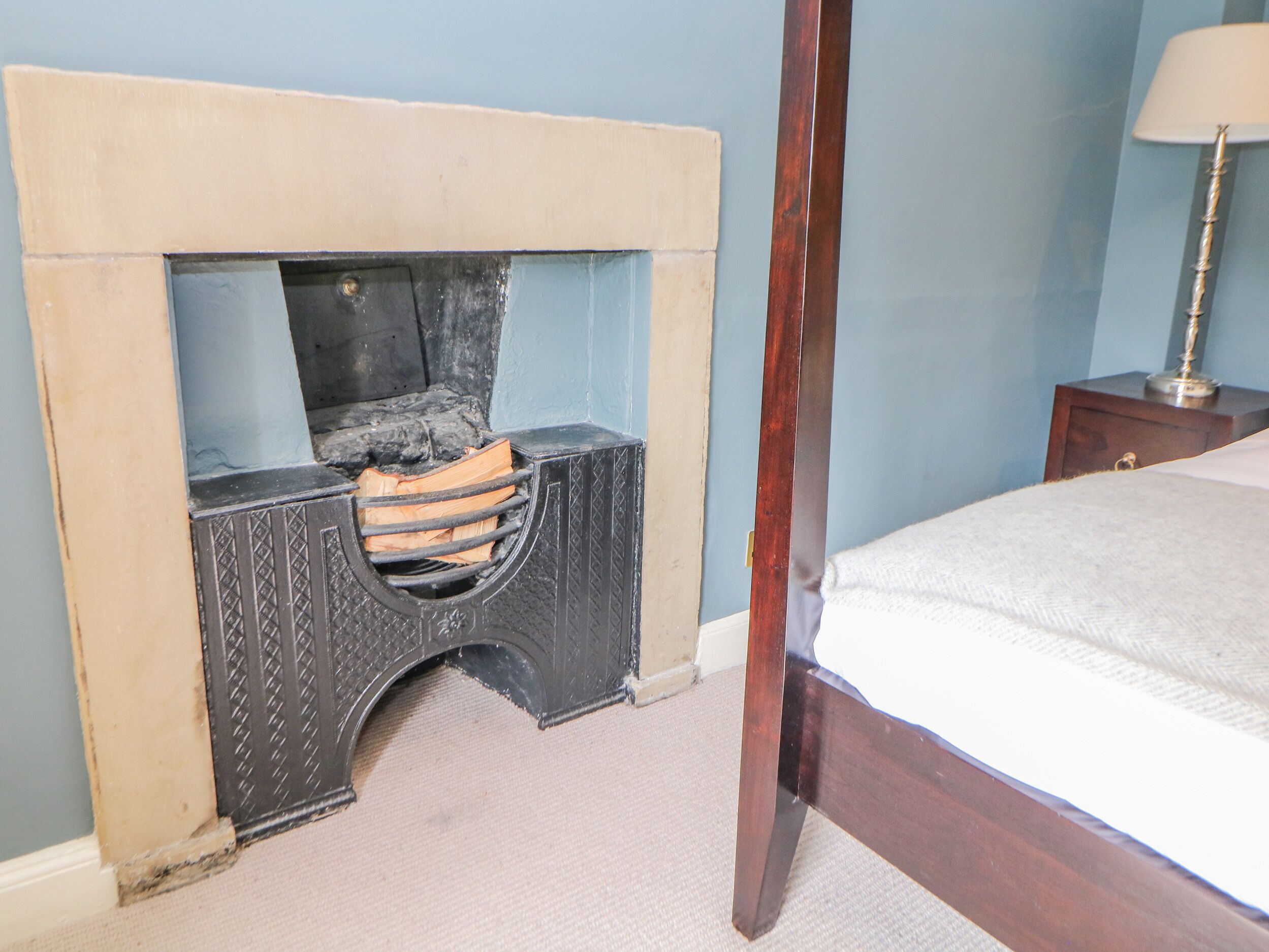 A bedroom with a bed, a nightstand, a lamp, and a fireplace with logs.