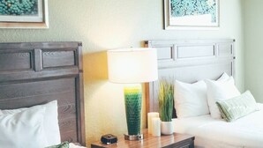 Ein Hotelzimmer mit einem hölzernen Kopfteil, einem Nachttisch mit Lampe, einer Topfpflanze und einem Bett mit weißer Bettwäsche.