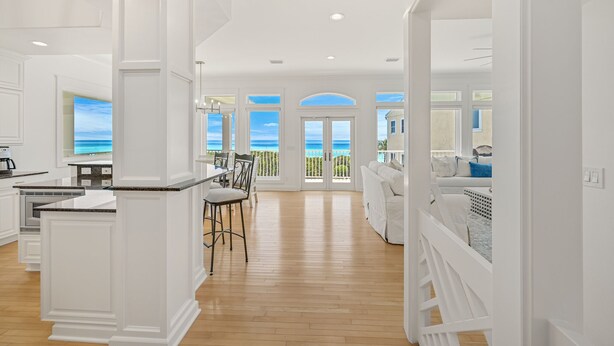 A spacious kitchen with white cabinetry and a large island, a dining area with a view of the ocean, and a living area with a white sofa and a balcony. - Gulf Front Updated Coastal Home 1 mile from Seaside (Santa Rosa Beach)