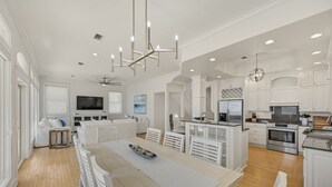 A spacious kitchen with white cabinetry, a dining area with a long table, and a living room with a sofa and TV. - Gulf Front Updated Coastal Home 1 mile from Seaside (Santa Rosa Beach)