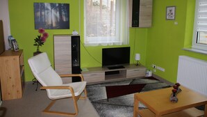 Un soggiorno con una parete verde, una sedia bianca, un tavolo di legno, una TV e una finestra con tenda bianca.