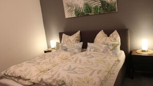 Un letto rifatto con cura, con una coperta a motivi fogliacei, due lampade sui comodini e una pianta appesa al muro.