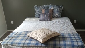 Un lit recouvert d’une couverture à carreaux et agrémenté de coussins décoratifs, un mur uni et un plancher en bois.