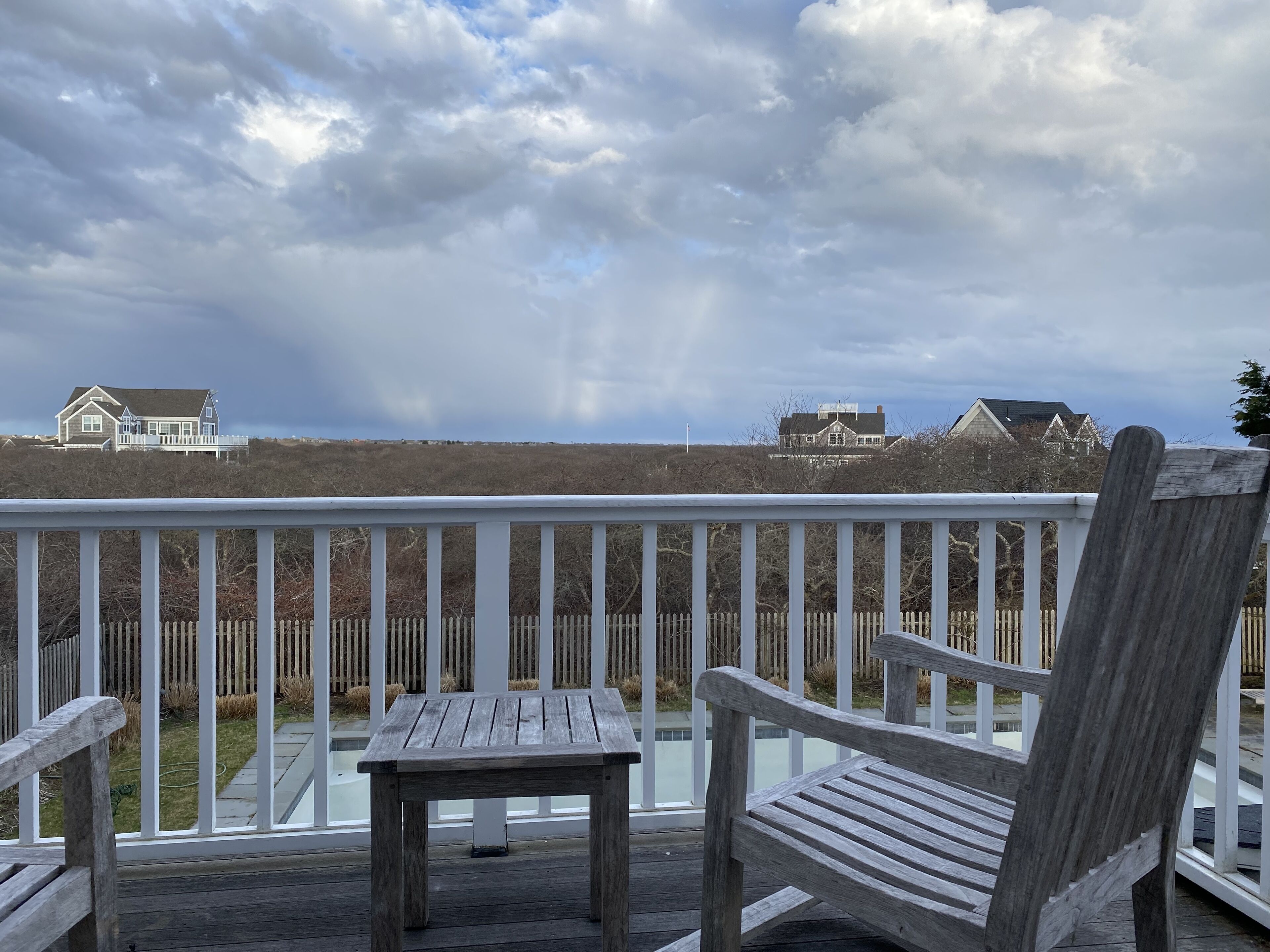 A wooden deck with two Adirondack chairs and a small table, overlooking a grassy area with houses and a cloudy sky.