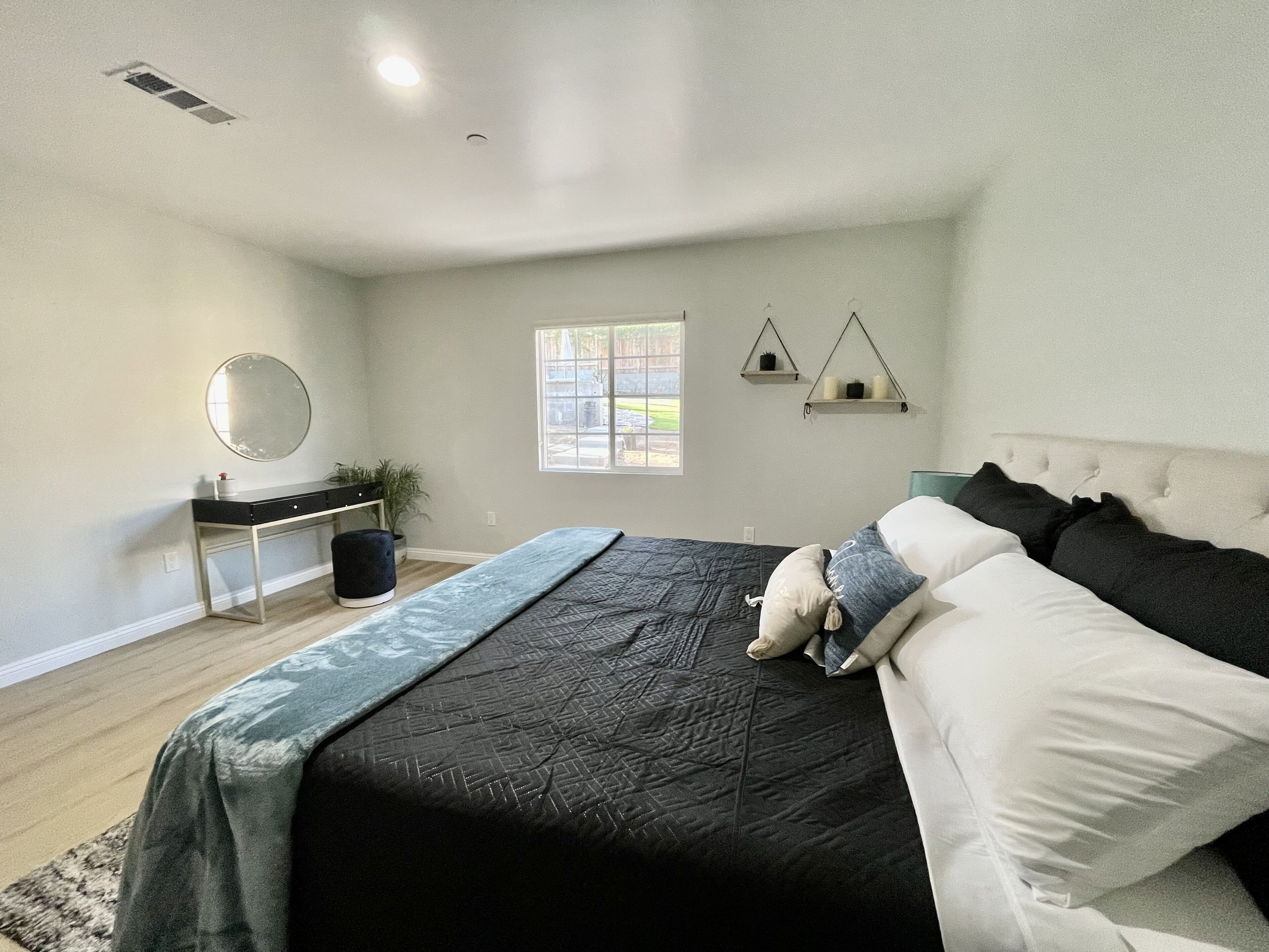 A bedroom with a bed, bedside table, mirror, window, and two wall-mounted shelves.