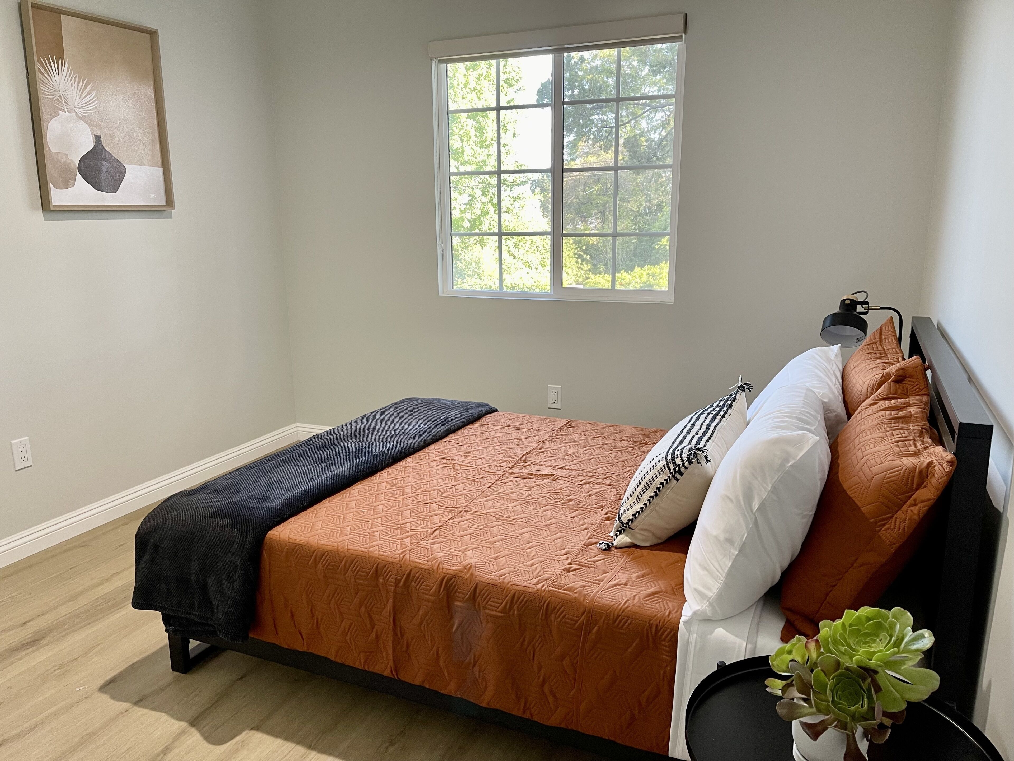 Un dormitorio con cama, ventana, lámpara de noche y una planta en maceta.
