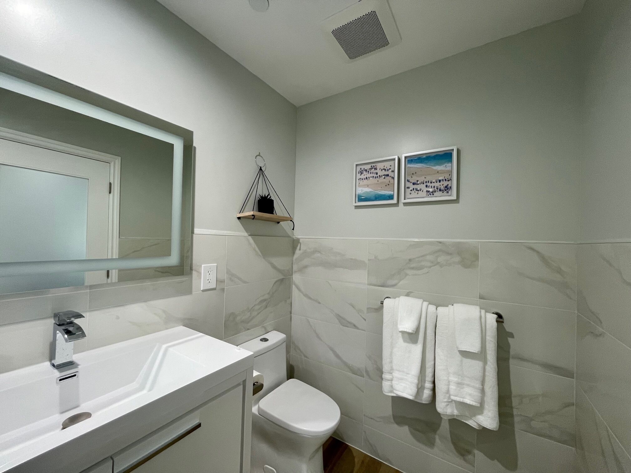 A bathroom with a white sink, toilet, and marble walls. There are two framed pictures on the wall and a towel rack with towels.