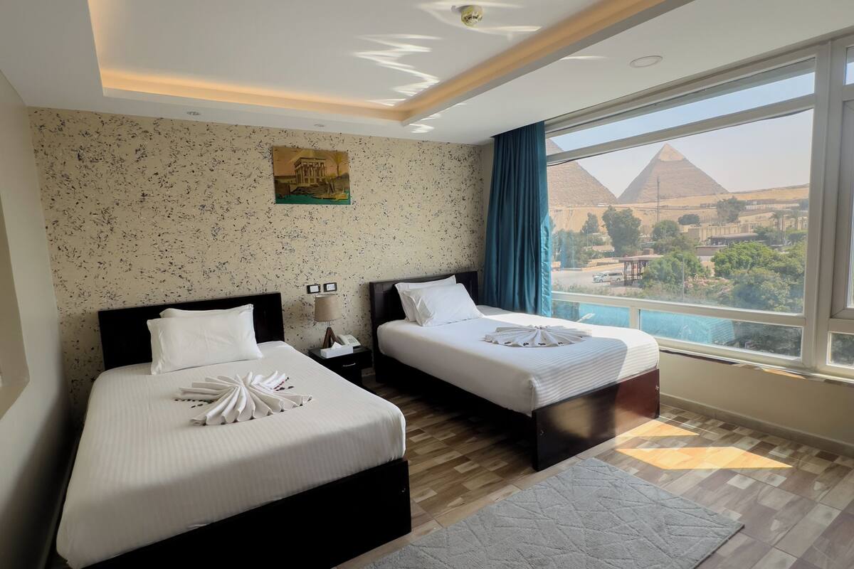 A hotel room with two beds, a large window with a view of pyramids, a wooden floor, and a painting on the wall.