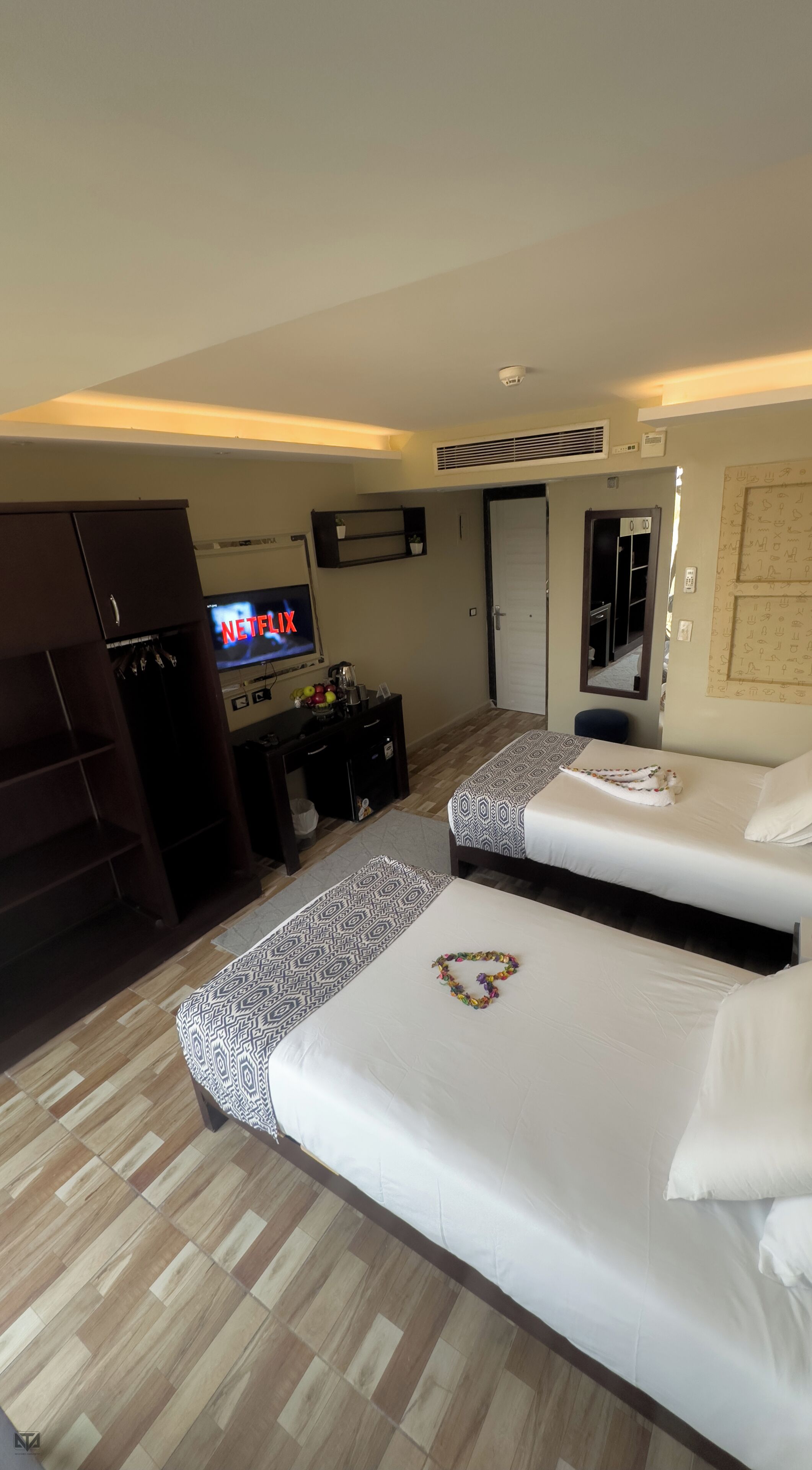 A hotel room with two beds, a TV displaying 'NETFLIX', a wardrobe, a desk with a computer, and a mirror.