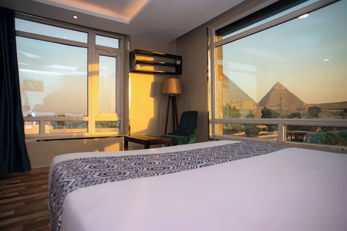 A hotel room with a large bed, a chair, a lamp, and a view of the pyramids.