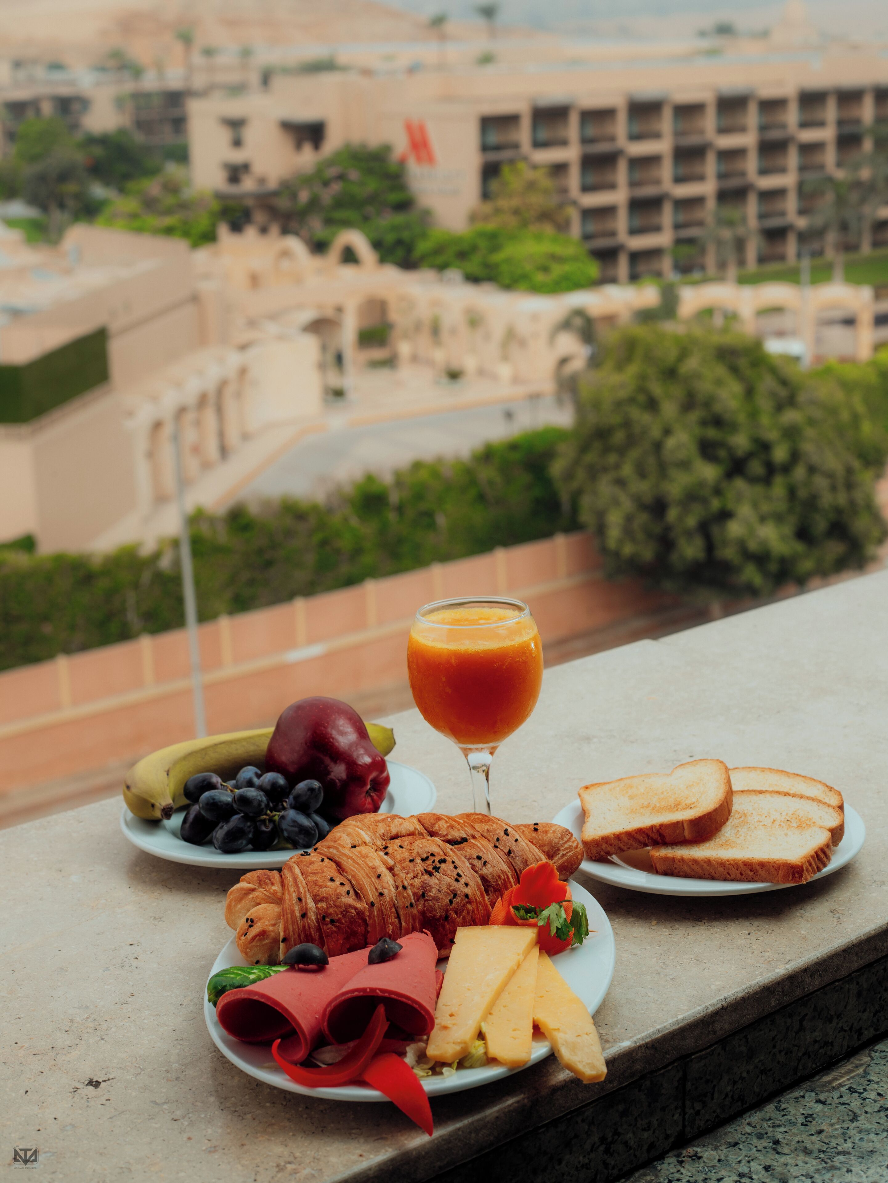 A meal with bread, cheese, ham, and vegetables on a plate, with a glass of orange juice, on a balcony overlooking a building with a flag.
