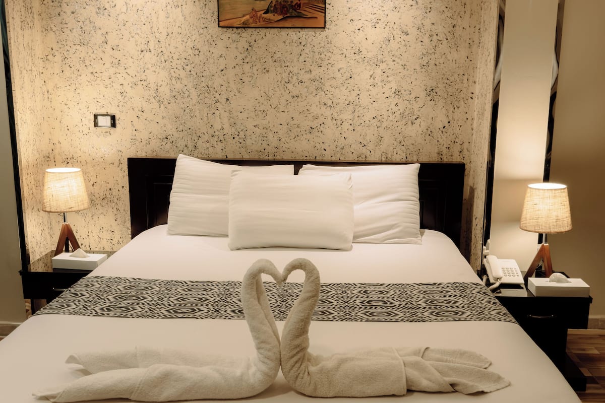 A hotel room with a large bed, two bedside lamps, a painting on the wall, and a heart-shaped towel arrangement on the bed.