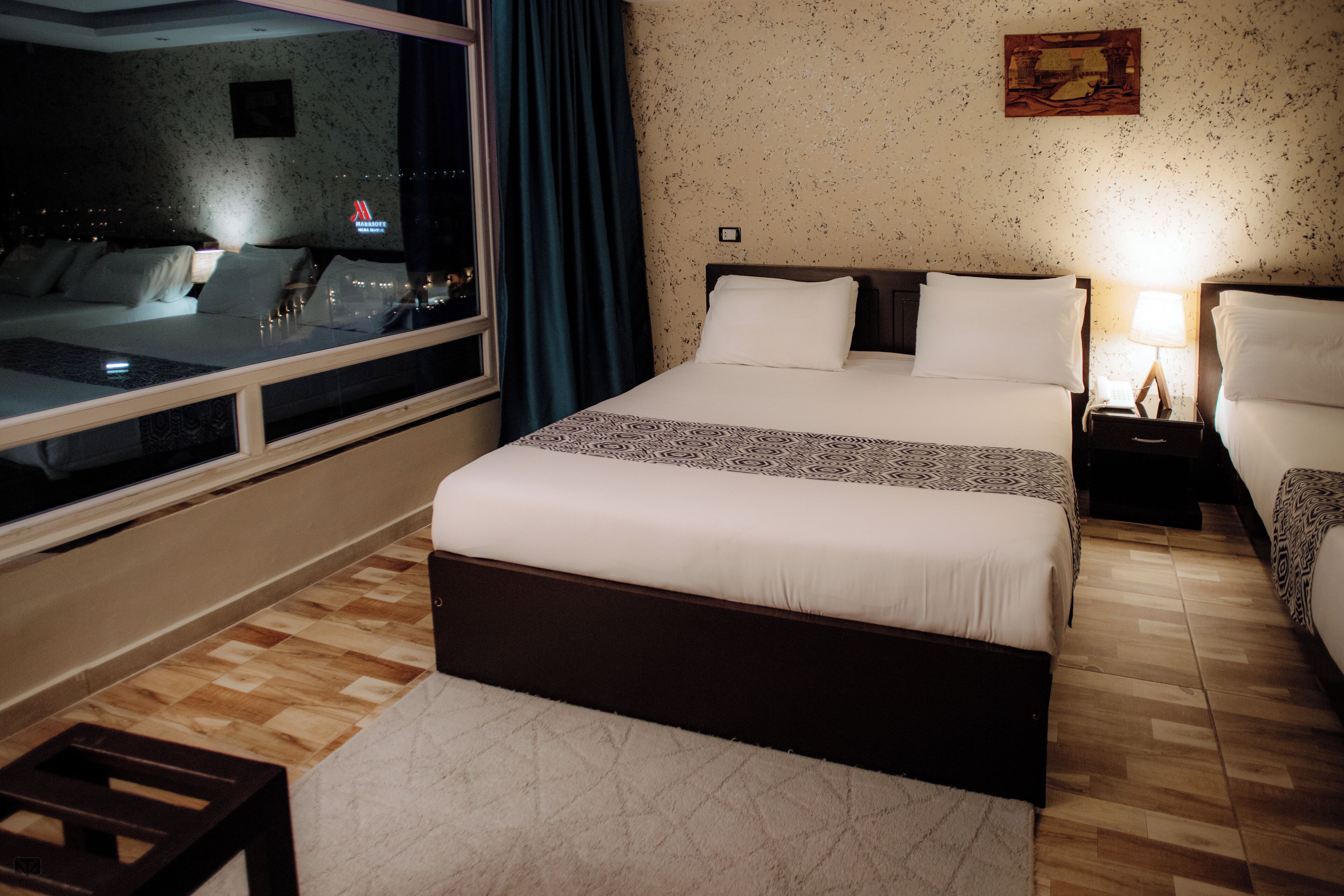 A hotel room with a large bed, a bedside table with a lamp, a window with a view of the city, and a wooden floor.