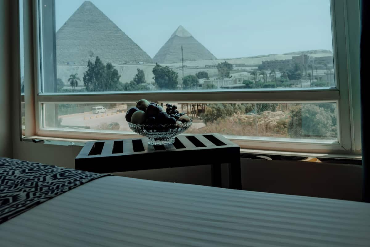 A room with a bed, a small table with a bowl of fruit, and a window with a view of pyramids.