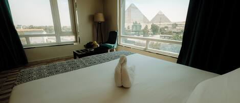 A hotel room with a large bed, a view of the pyramids, a chair, a lamp, and a small table with fruit.