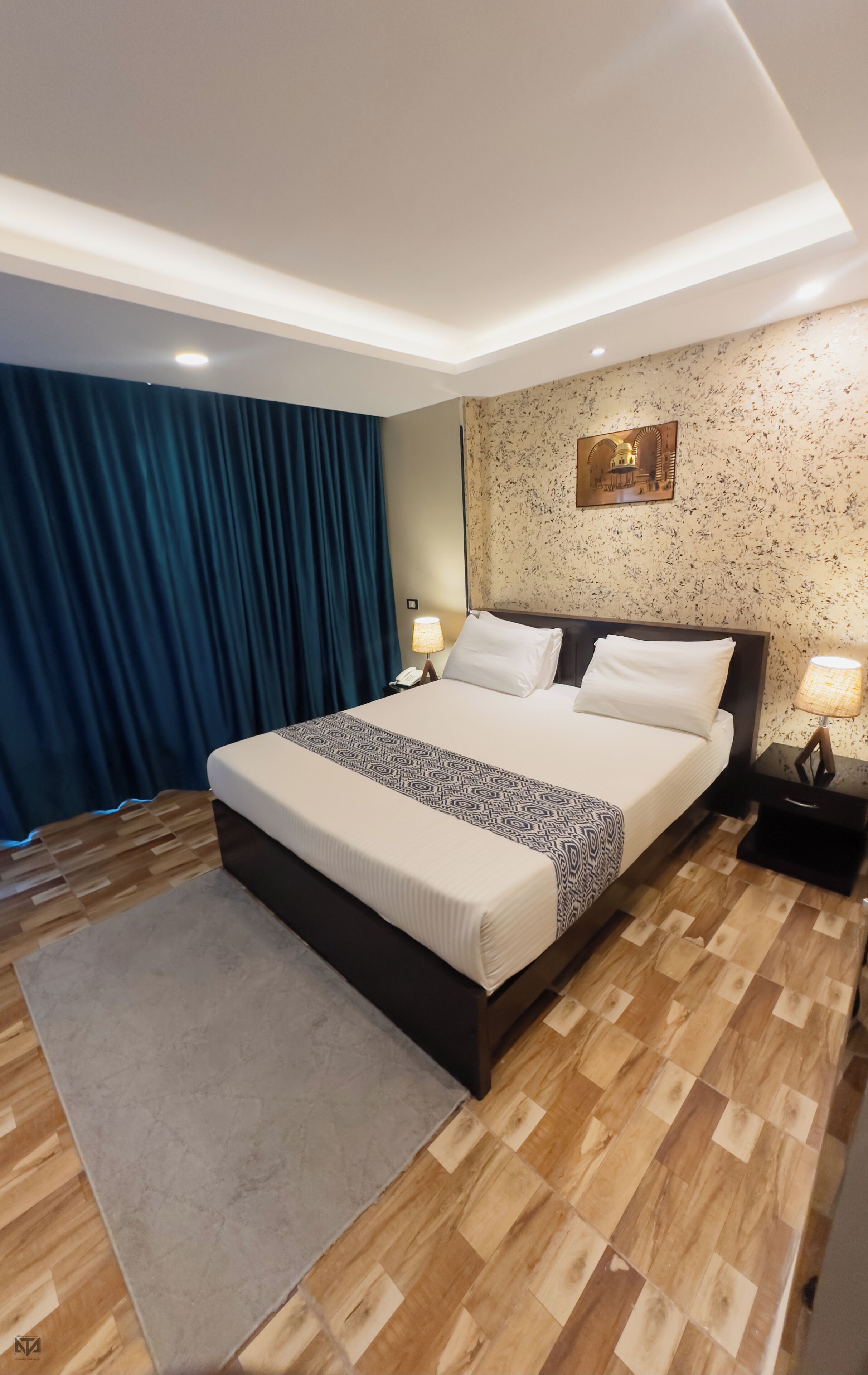 A modern hotel room with a large bed, a wooden floor, a blue curtain, and a wall-mounted lamp.