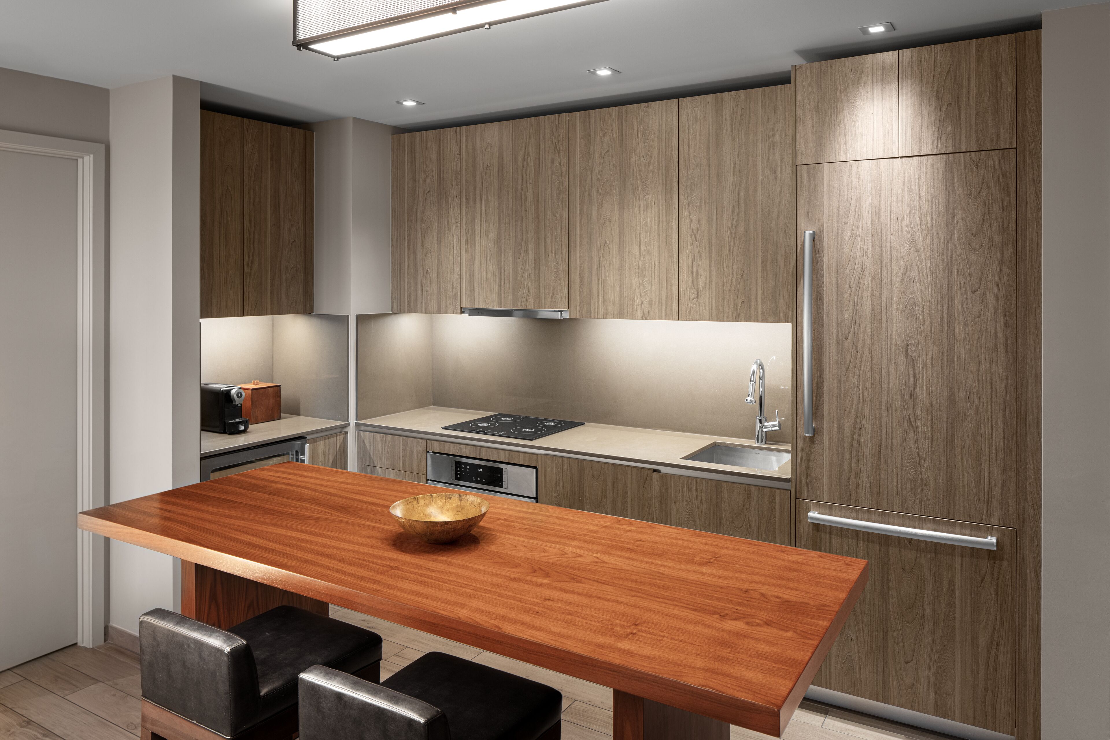 A modern kitchen with wooden cabinets, a stainless steel refrigerator, a built-in oven, and a wooden dining table with two chairs.