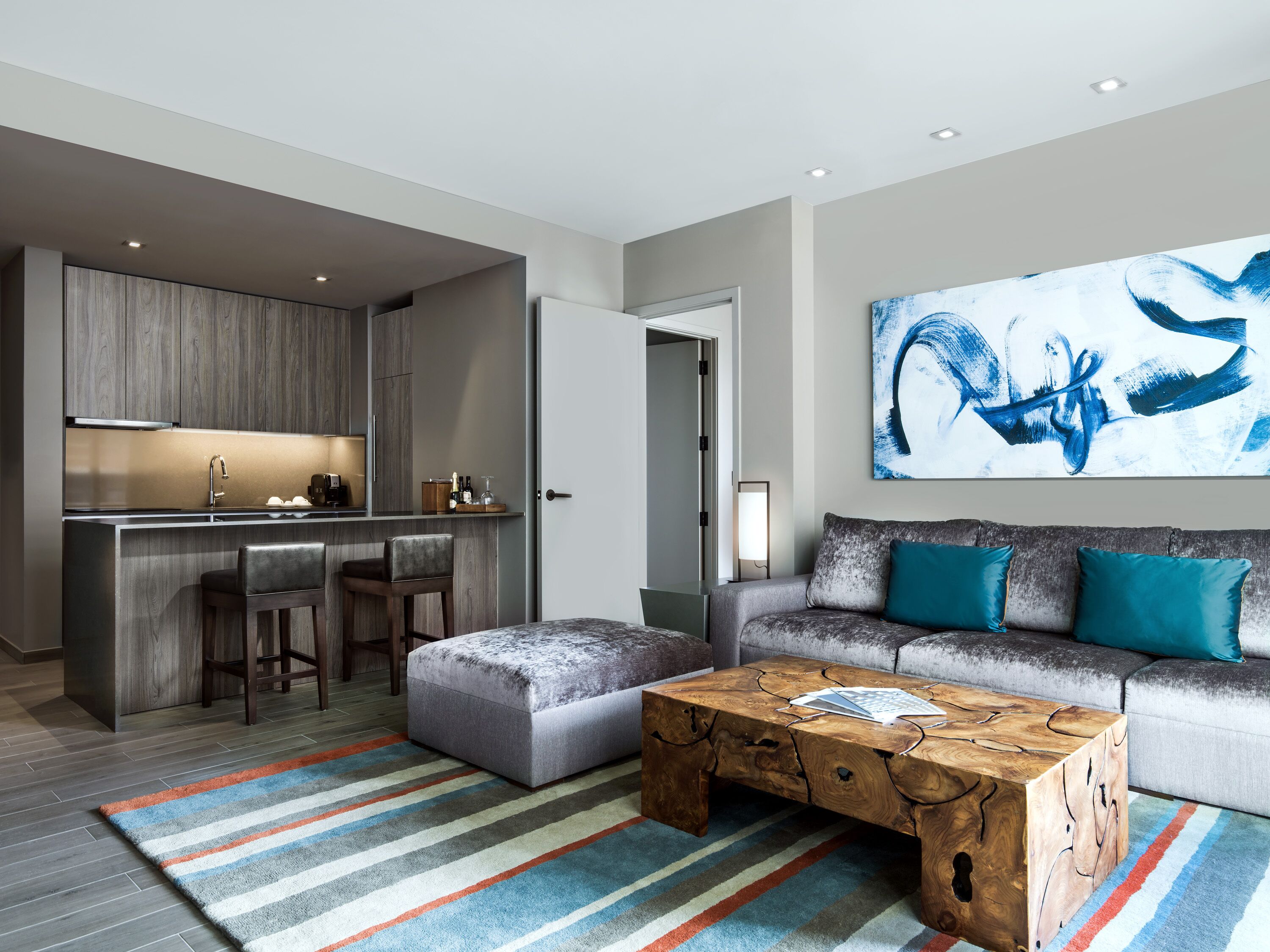 Ein modernes Wohnzimmer mit einem grauen Sofa, Türkis-farbenen Kissen, einem Holztisch und einem bunten Streifen Teppich.