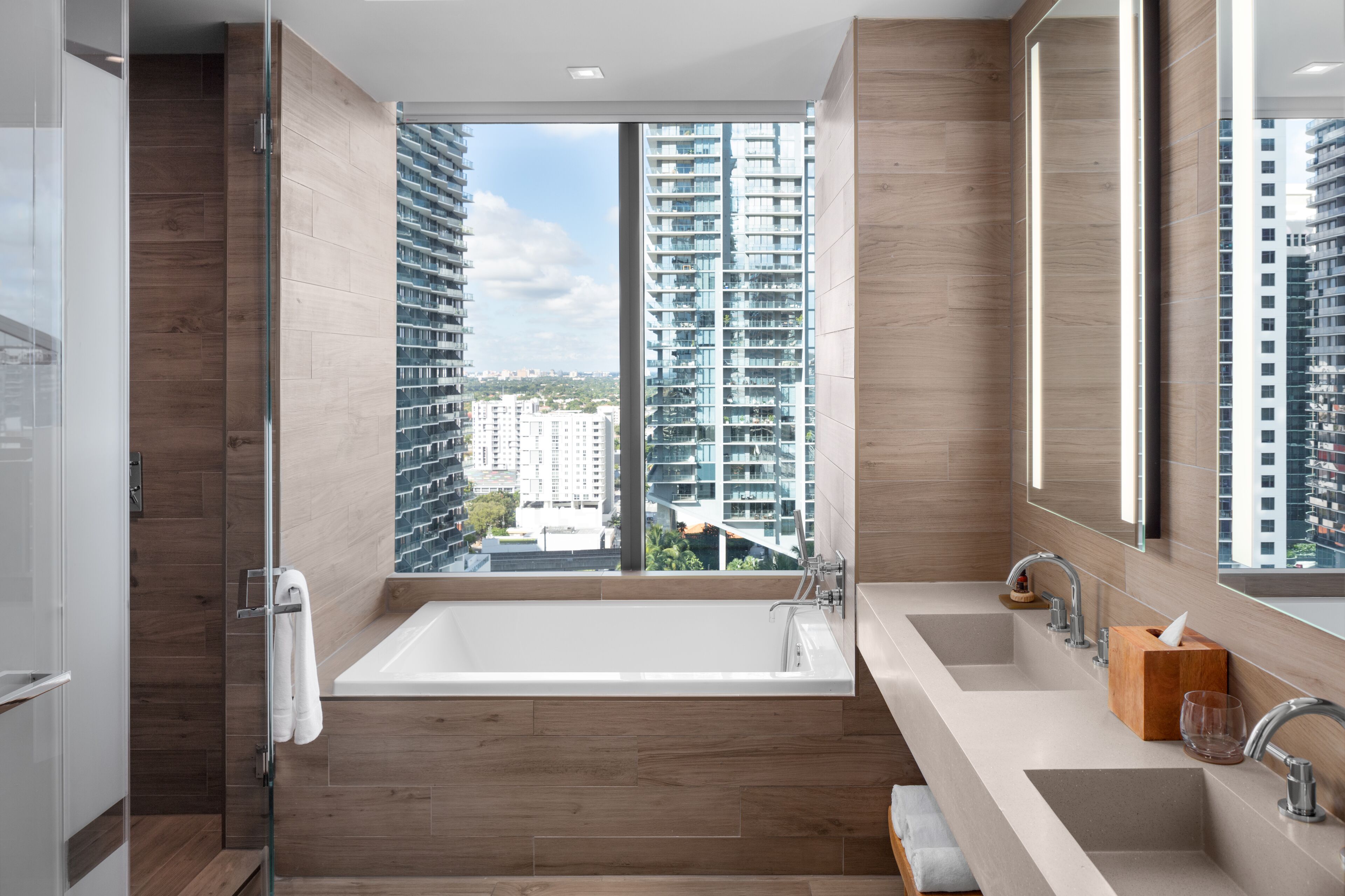 A modern bathroom with a large bathtub, dual sinks, and a large window offering a city view.