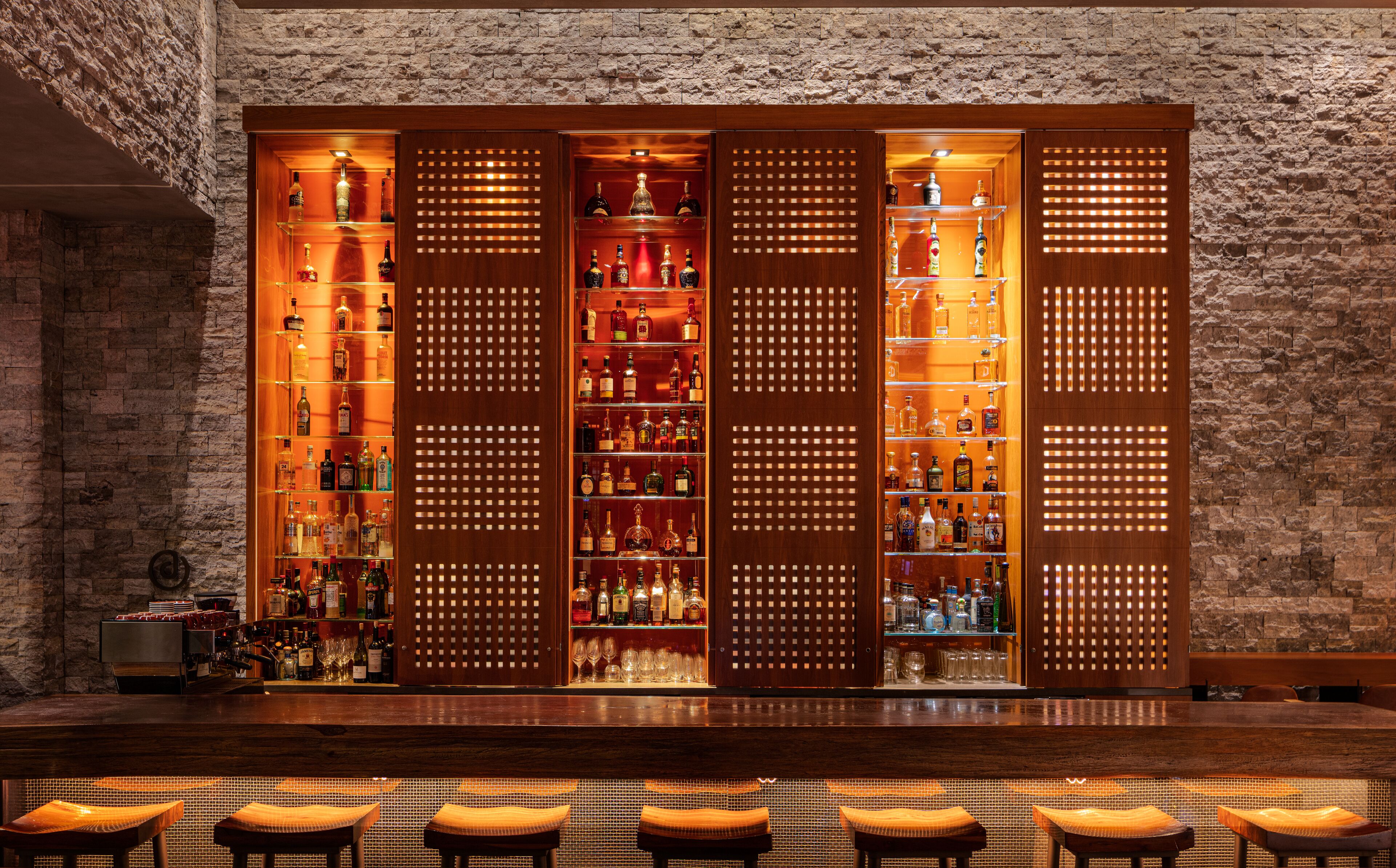 A bar area with a brick wall backdrop, wooden shelves stocked with bottles, and a seating area with cushioned stools.