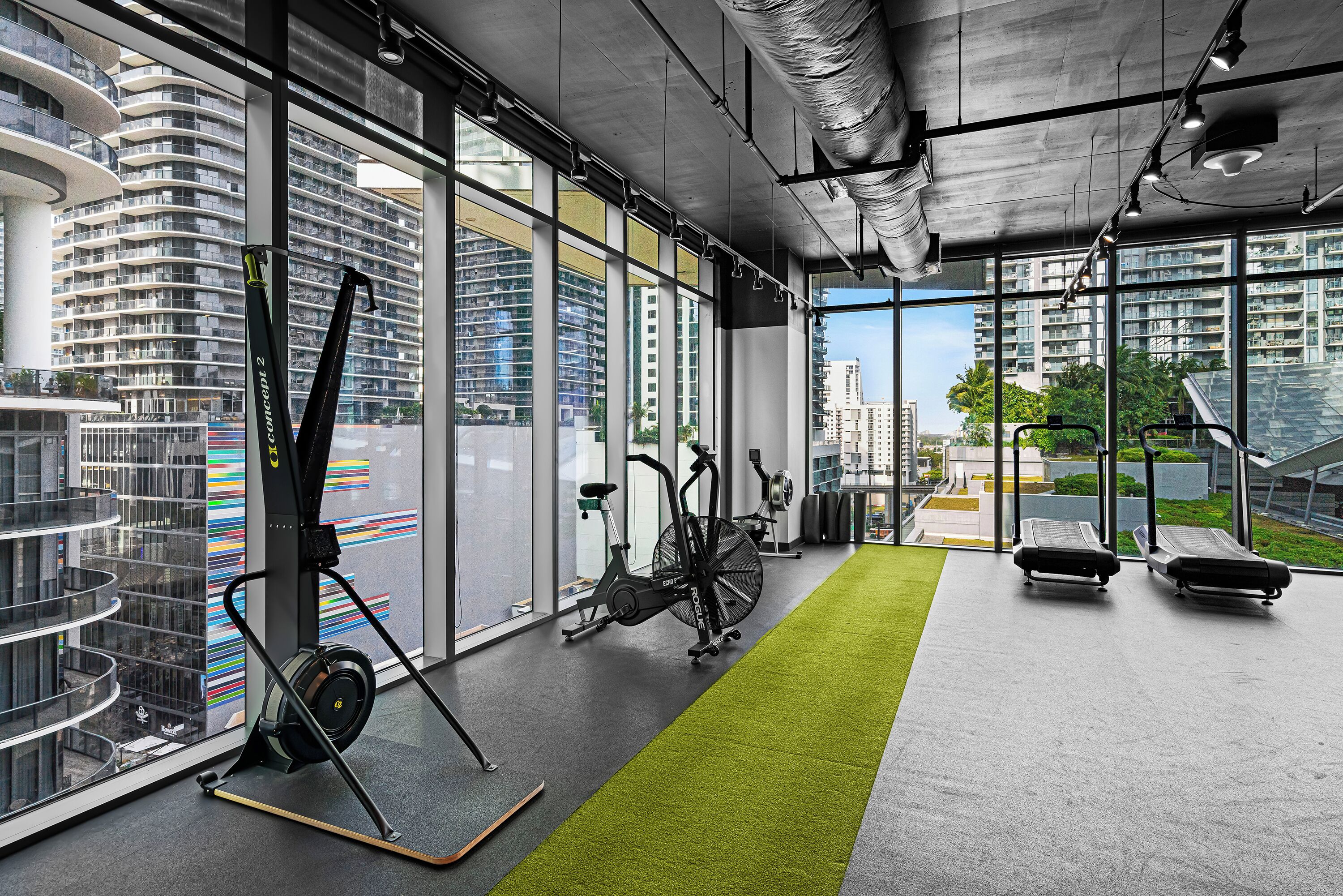 A modern gym with exercise equipment, large windows, and a view of high-rise buildings.