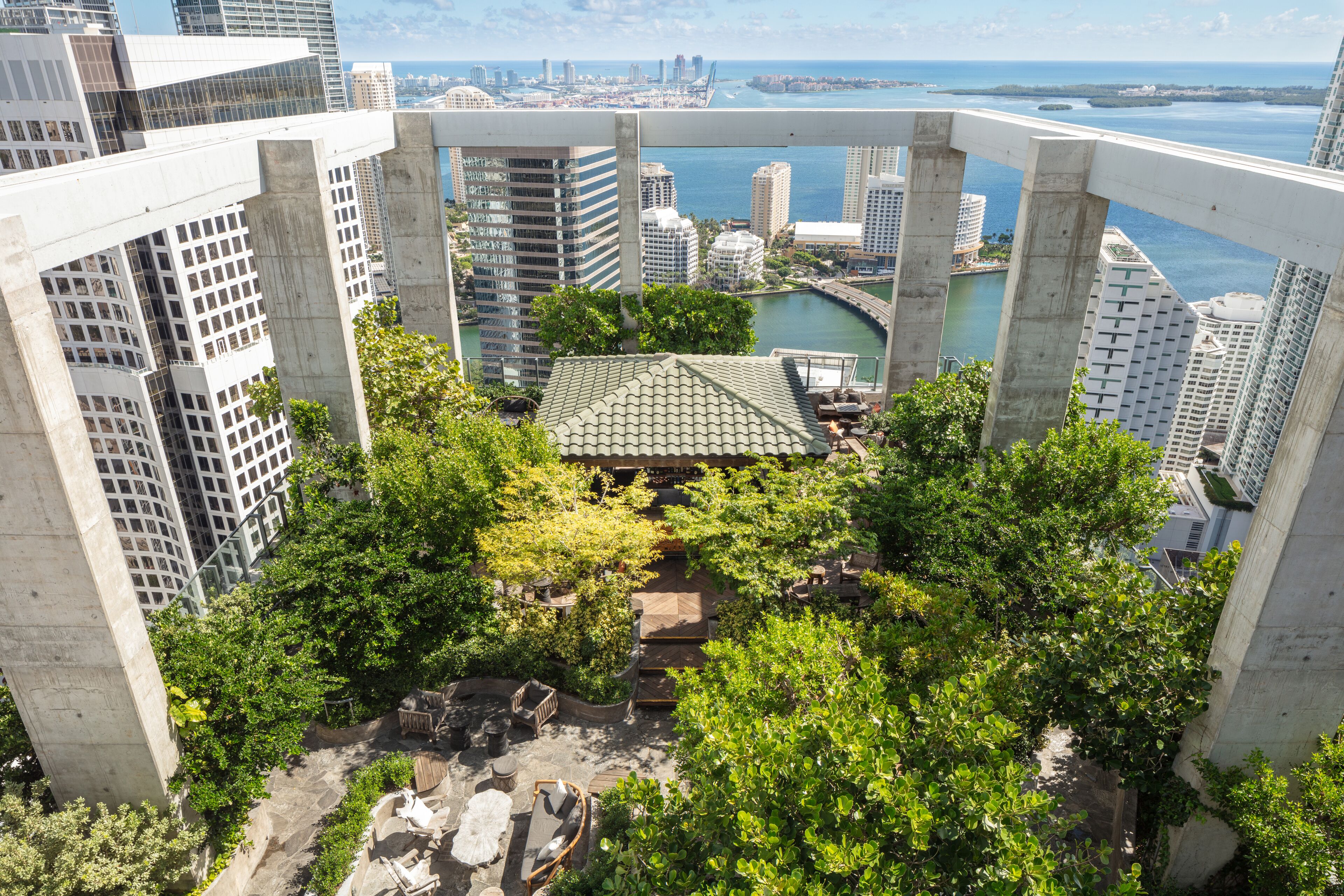 A high-rise building with a rooftop garden, surrounded by dense greenery and overlooking a cityscape and water body.