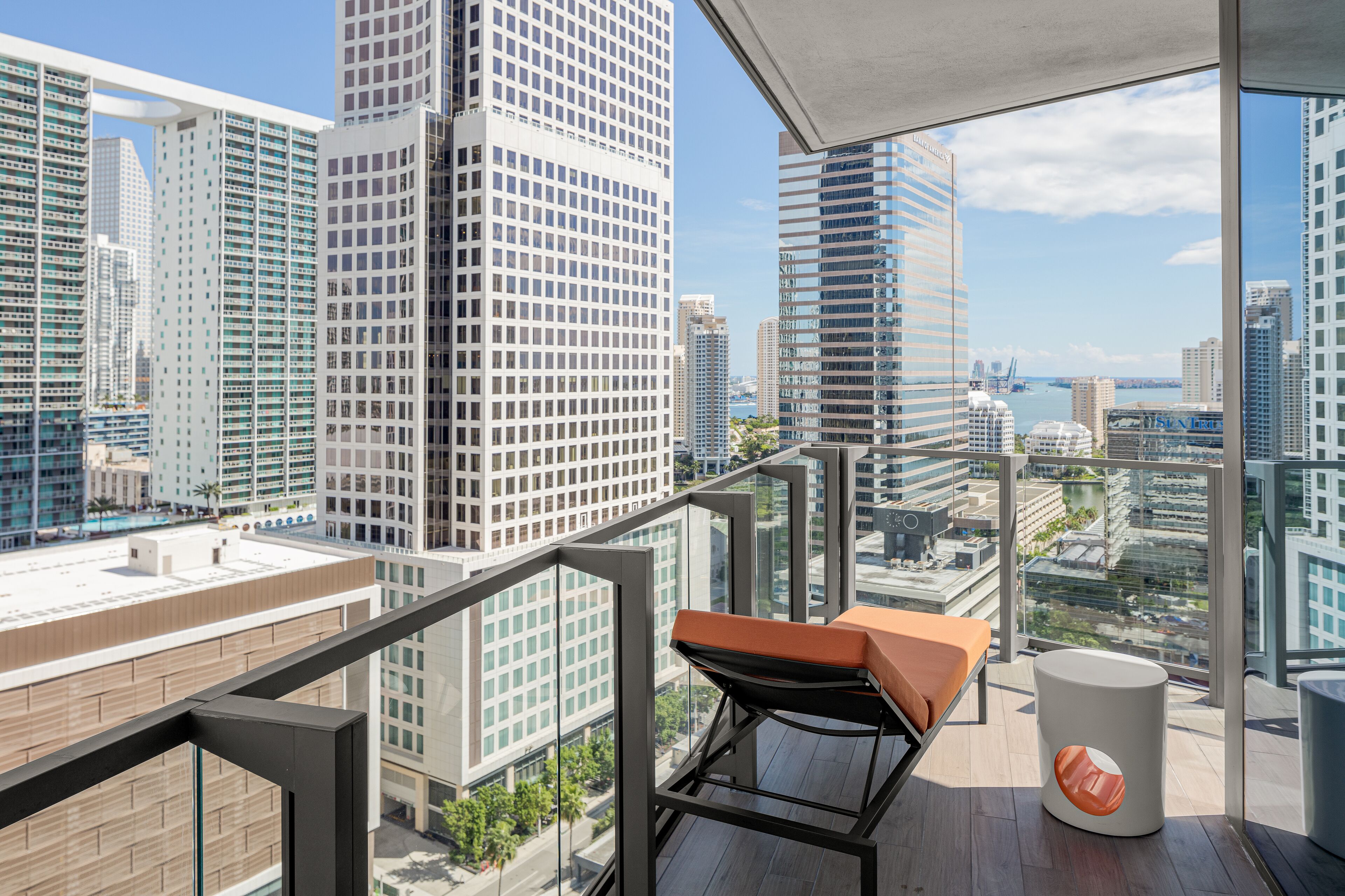 Un balcón en un edificio alto con una silla moderna y una pequeña mesa, que ofrece vistas de la ciudad y una masa de agua.