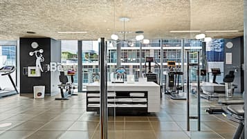 A modern gym with exercise equipment, a reception desk, and large windows offering views of the city.