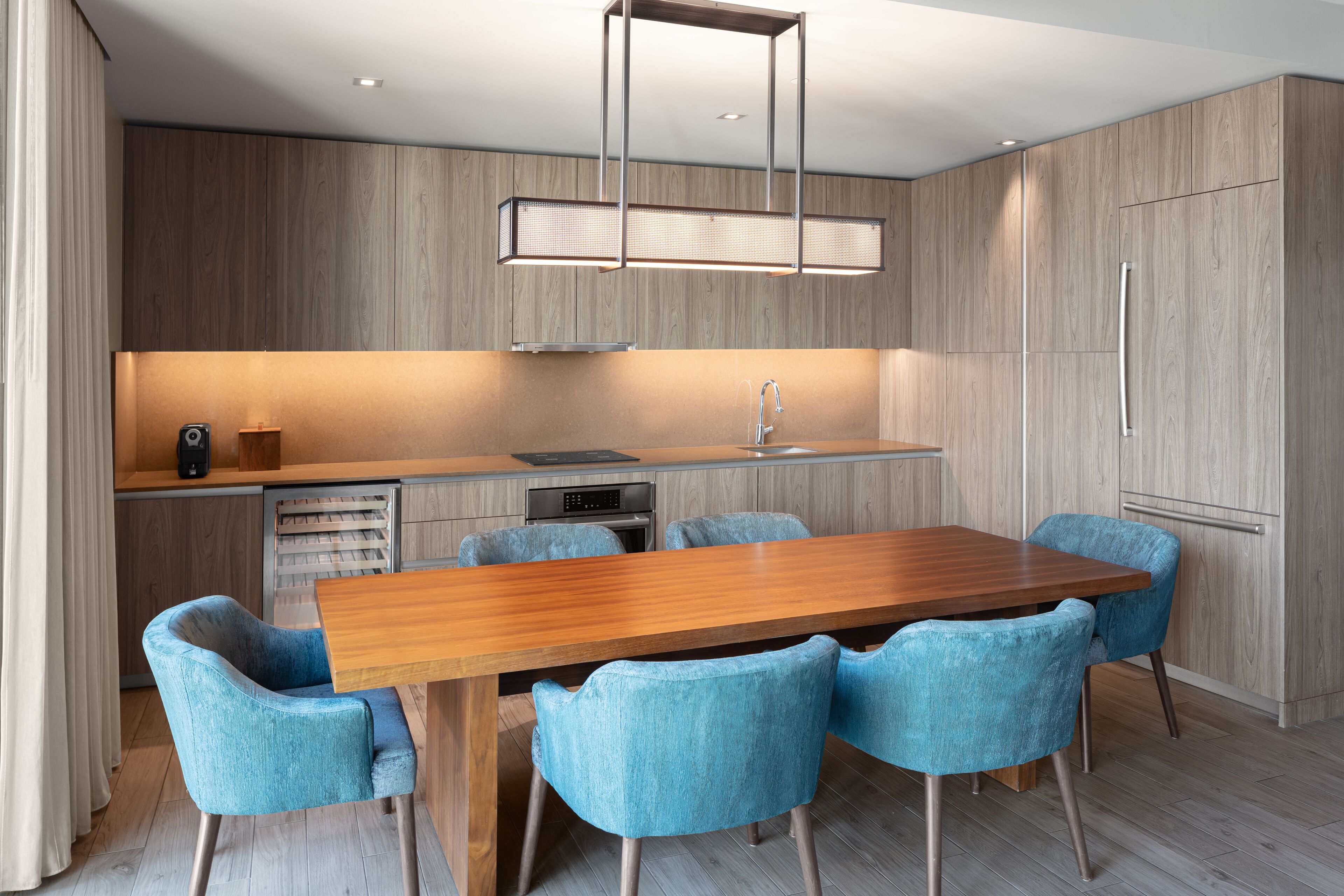 A modern kitchen with a wooden dining table and blue chairs, a built-in oven, and a stainless steel sink.