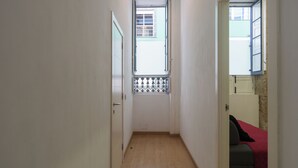 Un pasillo estrecho con suelo de madera, una puerta a la izquierda y una ventana con baranda de metal a la derecha.