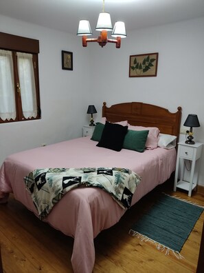 Un dormitorio con cama de madera, manta estampada en verde y blanco, dos lámparas de noche, mesita de noche de madera, ventana con cortinas y lámpara en el techo.