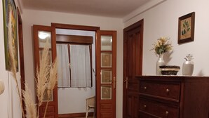 A room with wooden flooring, a wooden dresser, a small white ottoman, a green vase with dried plants, a chair, and a door leading to another room.