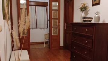 Een kamer met houten vloeren, een houten dressoir, een klein wit poefje, een groene vaas met gedroogde planten, een stoel en een deur naar een andere kamer.
