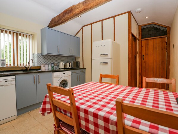 A compact kitchen with a washing machine, a microwave, and a refrigerator. There is a table with a checkered tablecloth and wooden chairs. - Sutton Barn (Ross-on-Wye)