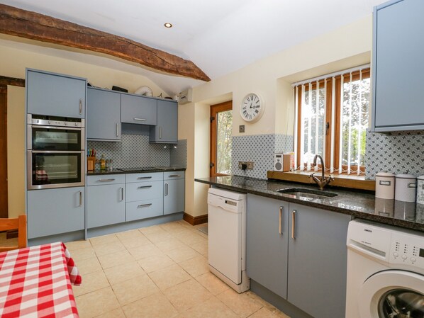 A compact kitchen with grey cabinets, a built-in oven, a dishwasher, and a window with curtains. - Sutton Barn (Ross-on-Wye)