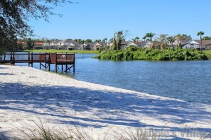 Une plage de sable avec une jetée en bois et des bancs, un lac paisible et des maisons en arrière-plan.