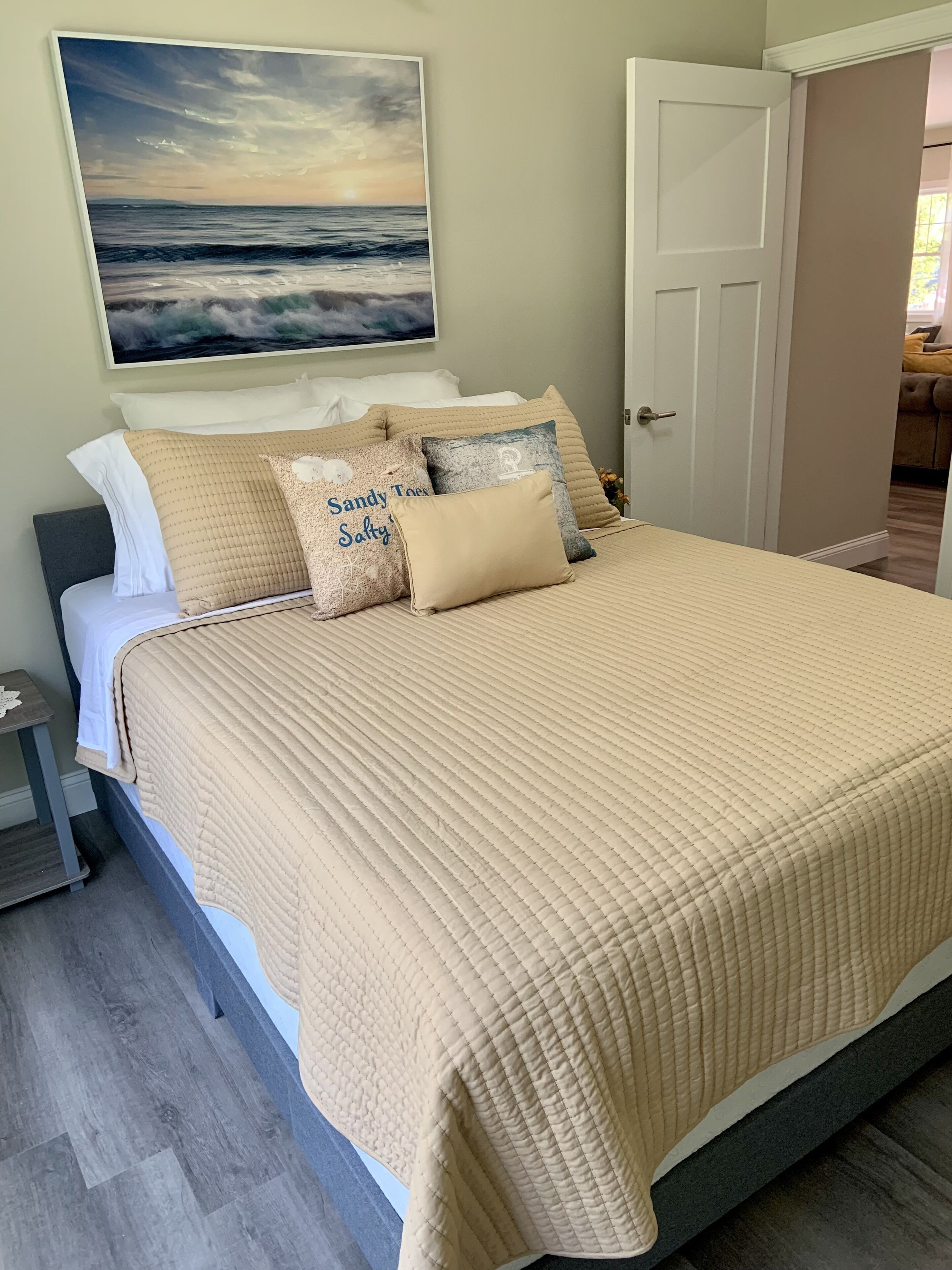 Une chambre à coucher avec un grand lit, une literie de couleur beige et un tableau représentant un coucher de soleil sur l’océan.
