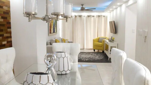 Una sala de estar moderna con una mesa de centro de vidrio, sofás blancos, un sillón amarillo, un ventilador de techo y un televisor de pantalla plana montado en la pared.