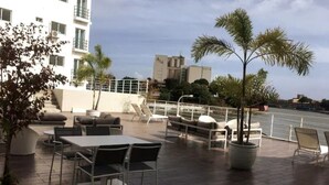 Un patio al aire libre con mesas y sillas, una palmera y vista al mar.